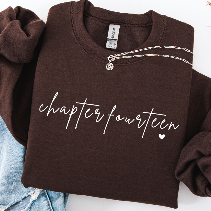 A folded dark brown sweatshirt with 'chapter fourteen' printed in white script across the front, styled with rolled sleeves and a silver chain necklace draped over the neckline. A small silver heart charm hangs from the chain.