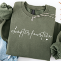 An olive green crewneck sweatshirt with 'chapter fourteen' printed in white script across the chest, styled flat-lay with rolled sleeves and a silver necklace on a white background.