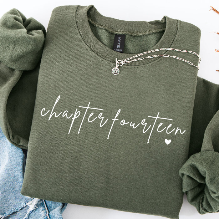 An olive green crewneck sweatshirt with 'chapter fourteen' printed in white script across the chest, styled flat-lay with rolled sleeves and a silver necklace on a white background.